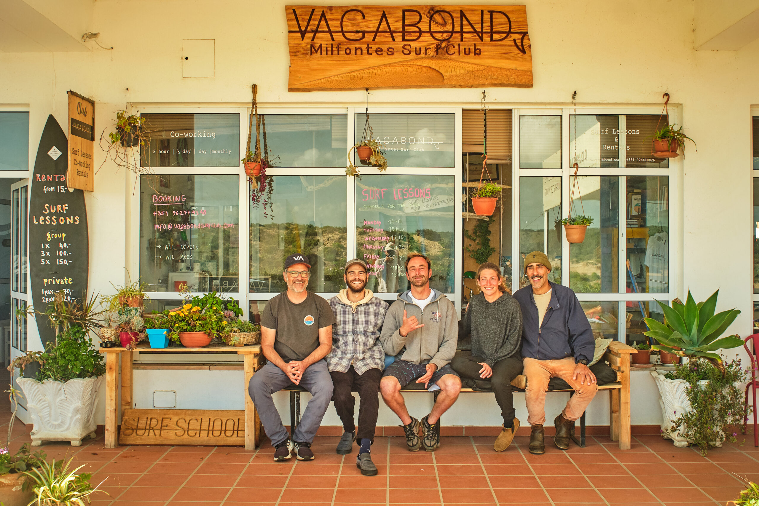 Club Vagabond surf instructor team sitting together on a bench in front of the shop, all smiling at the camera.