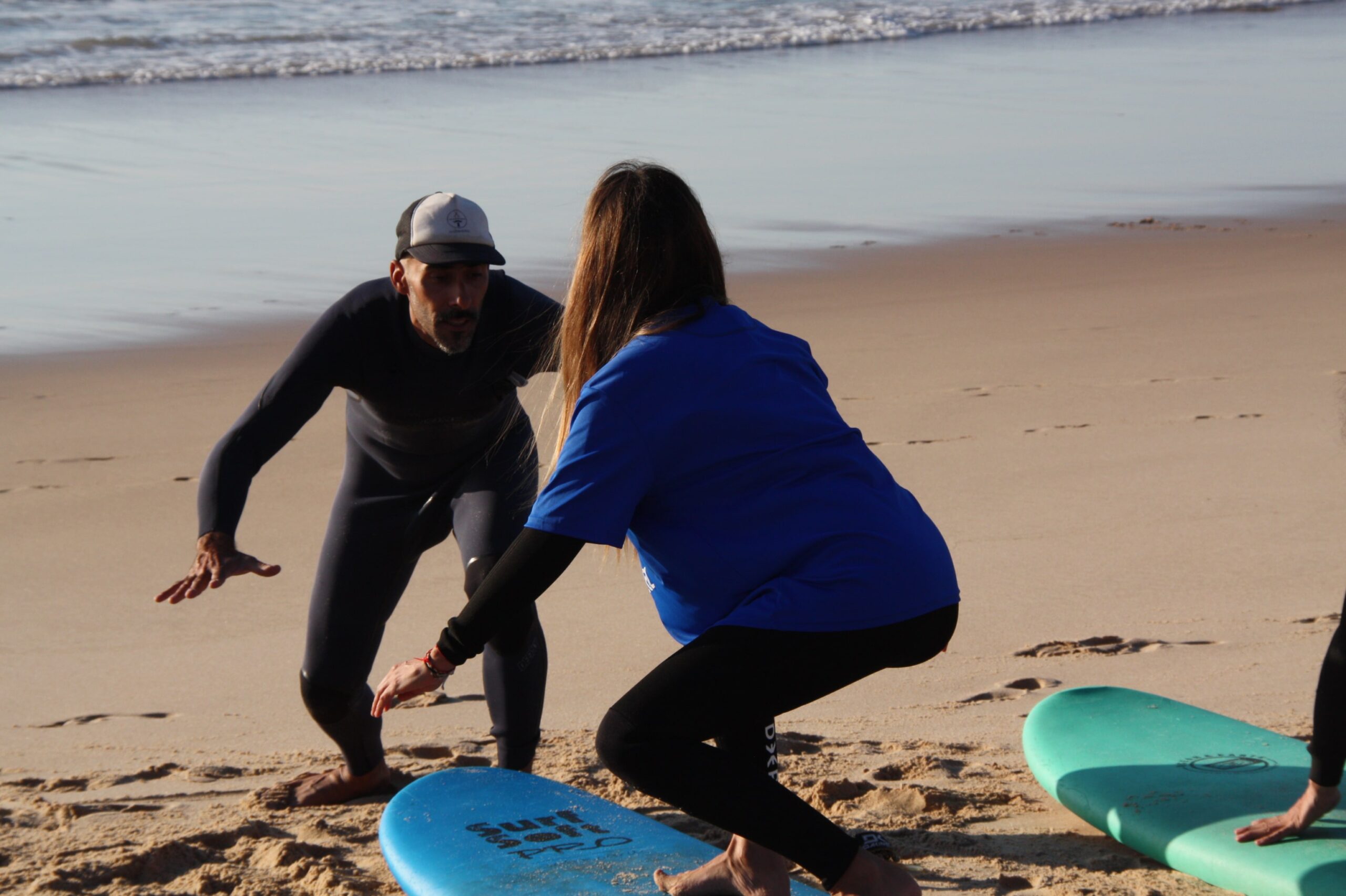 A student practicing the correct stance for surfing on the sand during a beginner lesson, mirroring surf instructor Ivo as he demonstrates the posture.