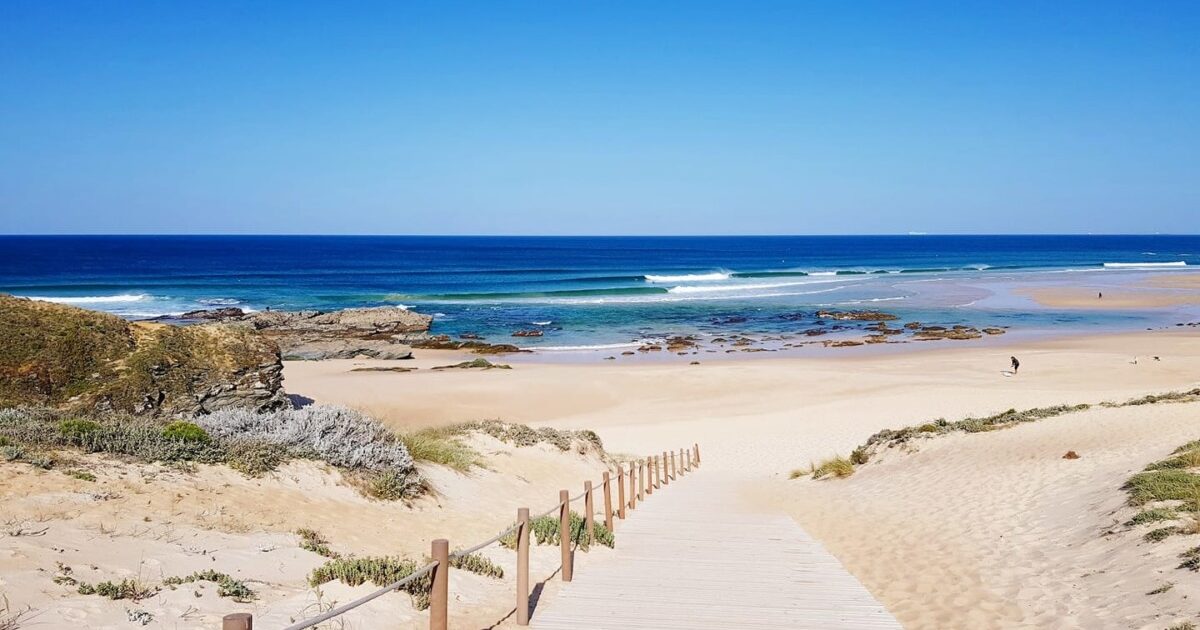 Sandy downhill path at Malhão Beach opening onto a wide sandy shore with gentle waves rolling in.