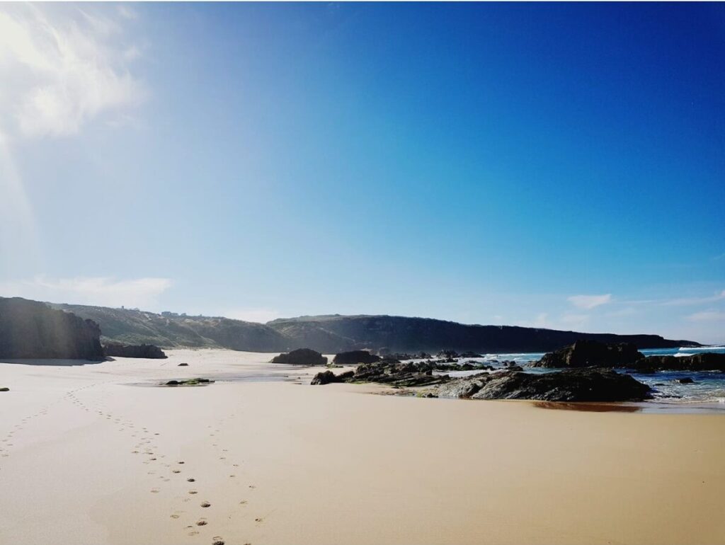 Wide sandy Furnas Beach with footprints in the sand, rocks scattered in the water, and shadows cast on the shore by nearby cliffs.