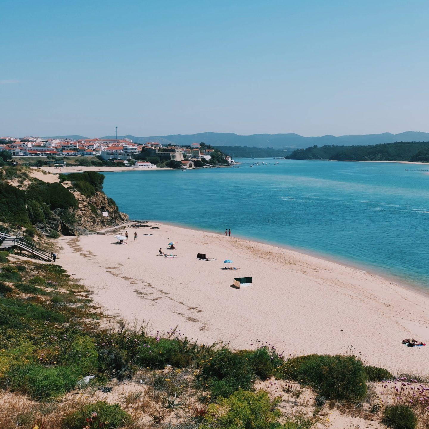 Scenic view of Vila Nova de Milfontes and the Mira River under a clear blue sky, with a sandy beach lining the riverbank.