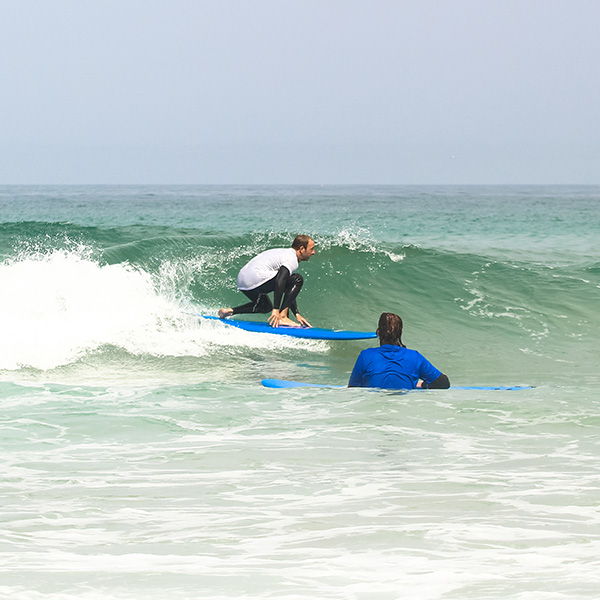Surf instructor Pedro riding a wave, demonstrating correct surfing posture and technique, while a surf student watches attentively from the water.