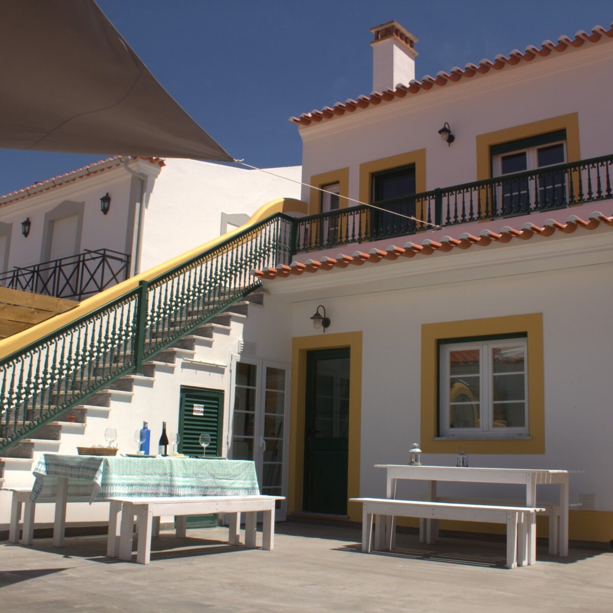 Sunlit outdoor area of Retromovement House with benches and tables, a two-storey building in view, and stairs leading up to one of the apartments.
