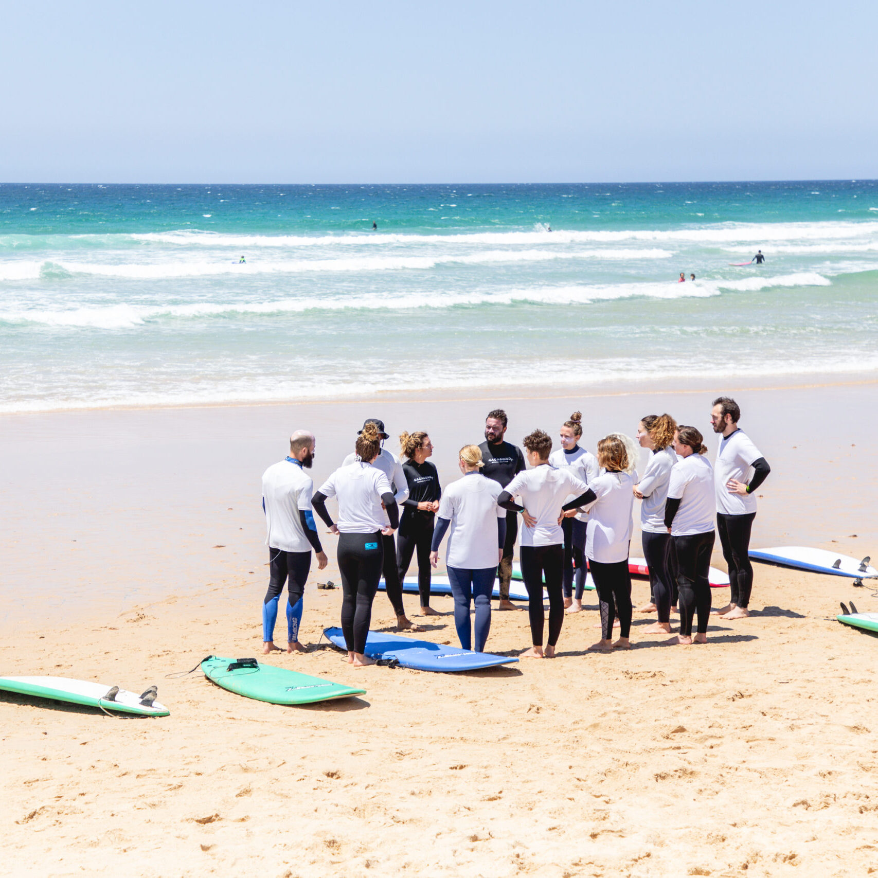 Group of surf students standing in a circle with two Club Vagabond instructors on the sandy beach of Furnas on a sunny day.