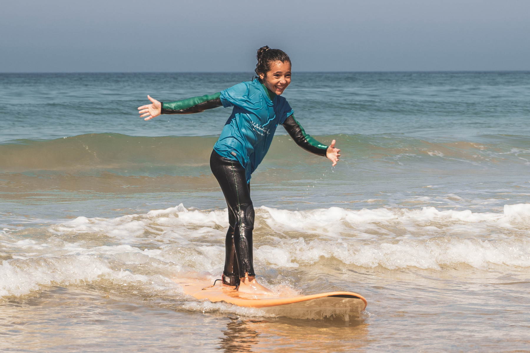 Girl standing and smiling on a surfboard riding a small wave on a sunny day.