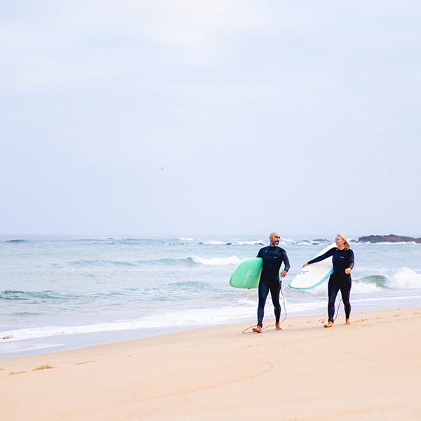 Surf instructor Ivo walking along the beach with a surf student, both smiling and talking while carrying their surfboards.