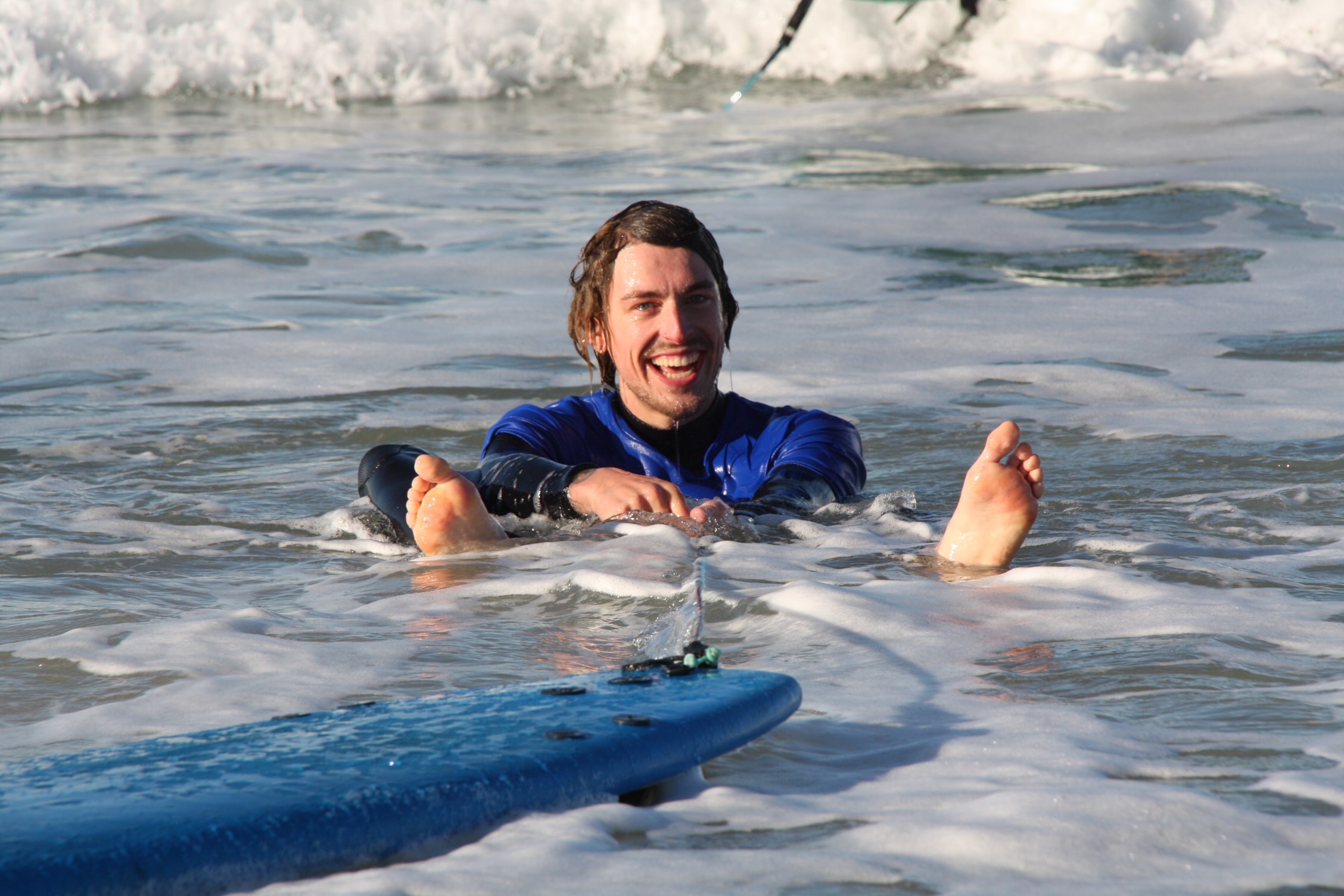 Surf student with Club Vagabond shirt sitting in the water, smiling, with leash in between his legs and the surf board floating in front of him.