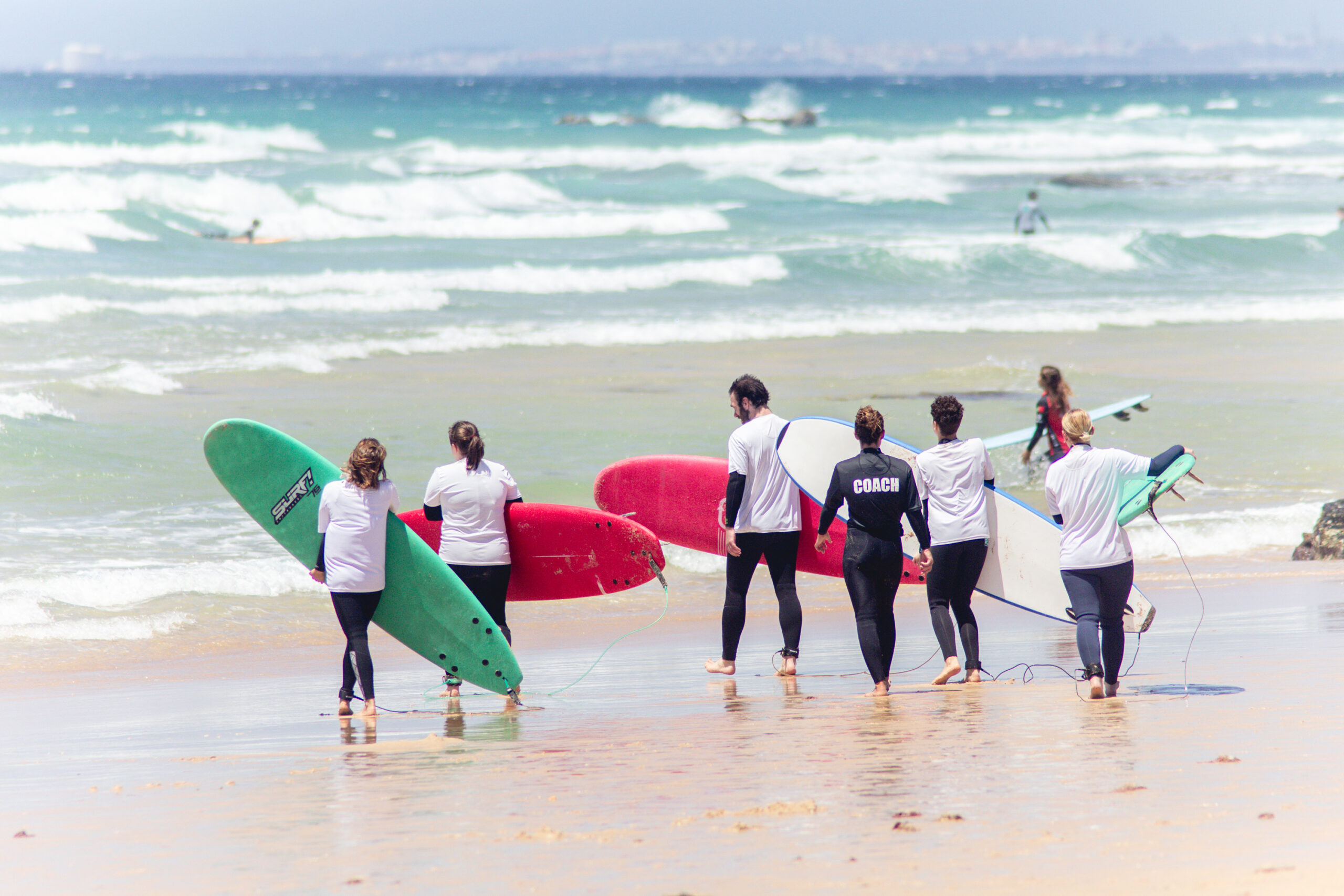 Surf students walking toward the waves with a Club Vagabond surf instructor, all carrying surfboards and ready to begin their lesson.