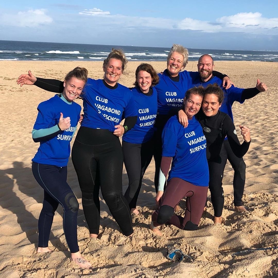 Group of surfers wearing blue Surf Club Vagabond shirts and wetsuits standing on the beach, ready to begin a surf lesson.