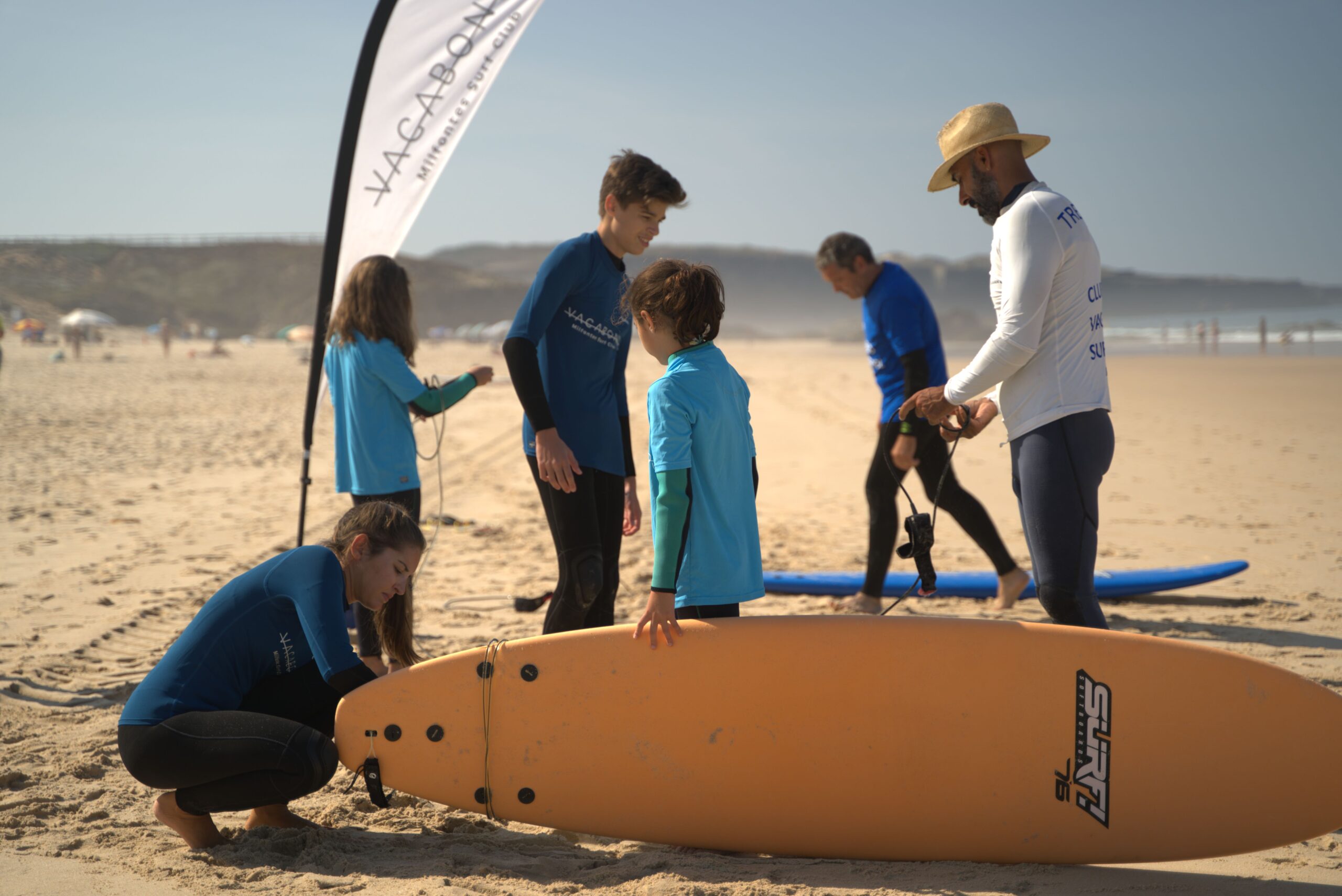 Surf instructor Ivo helping a student by opening the leash of a surfboard, while other students prepare their boards on the beach; a Vagabond flag is planted in the sand, fluttering in the wind.