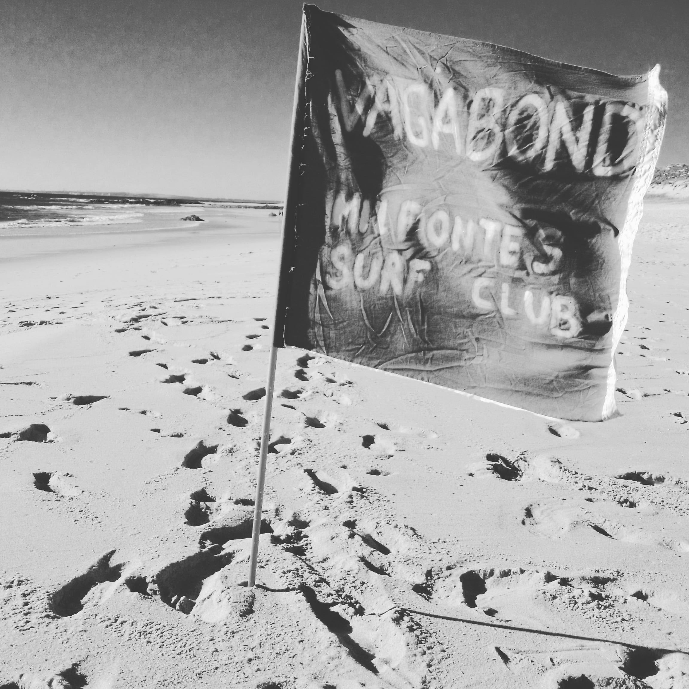 Vagabond Surf Club flag blowing in the wind at Furnas Beach, Vila Nova de Milfontes, with golden sand, gentle waves, and a clear blue sky in the background.