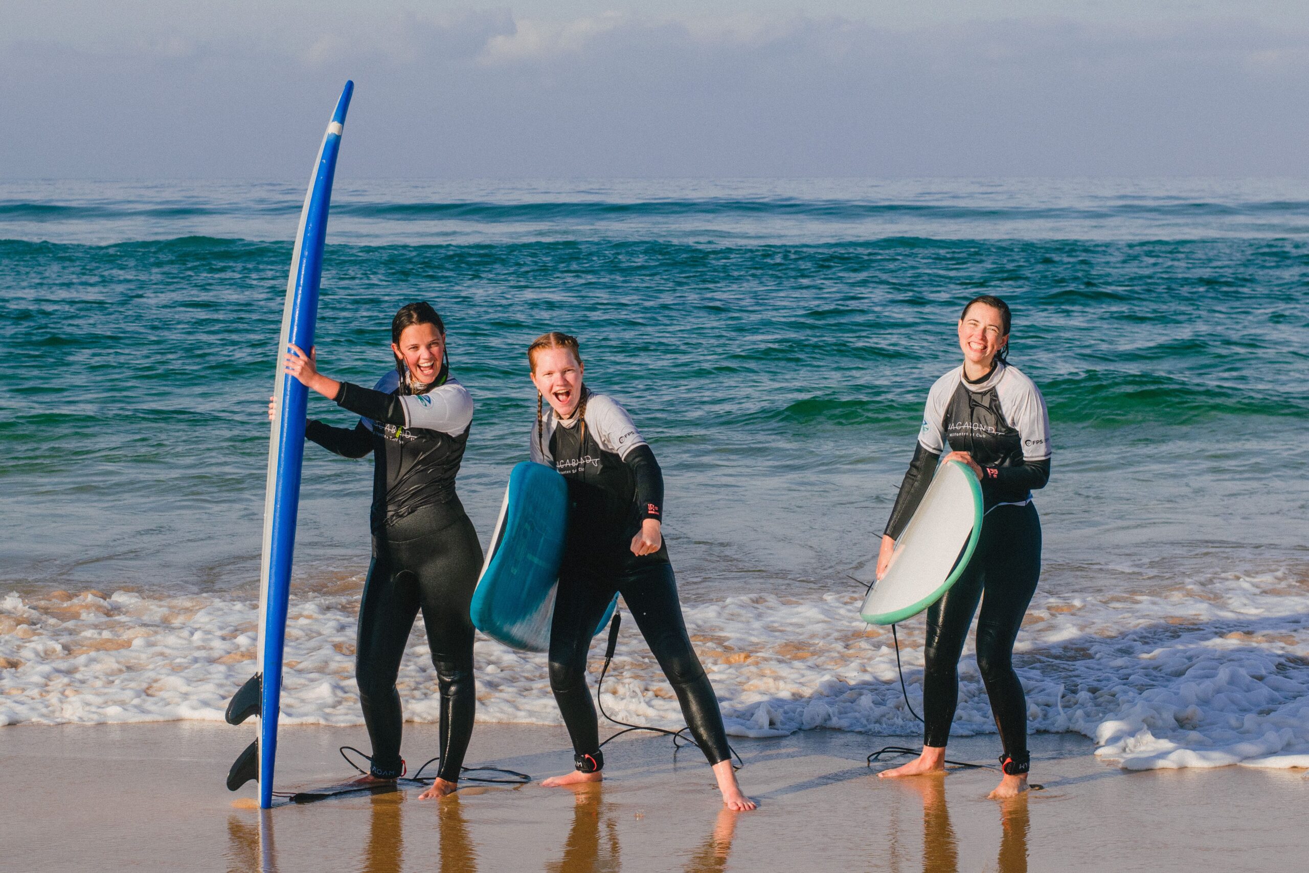 Three smiling girls walking out of the water with their surfboards in hand, cheerful and full of energy after a surf session.