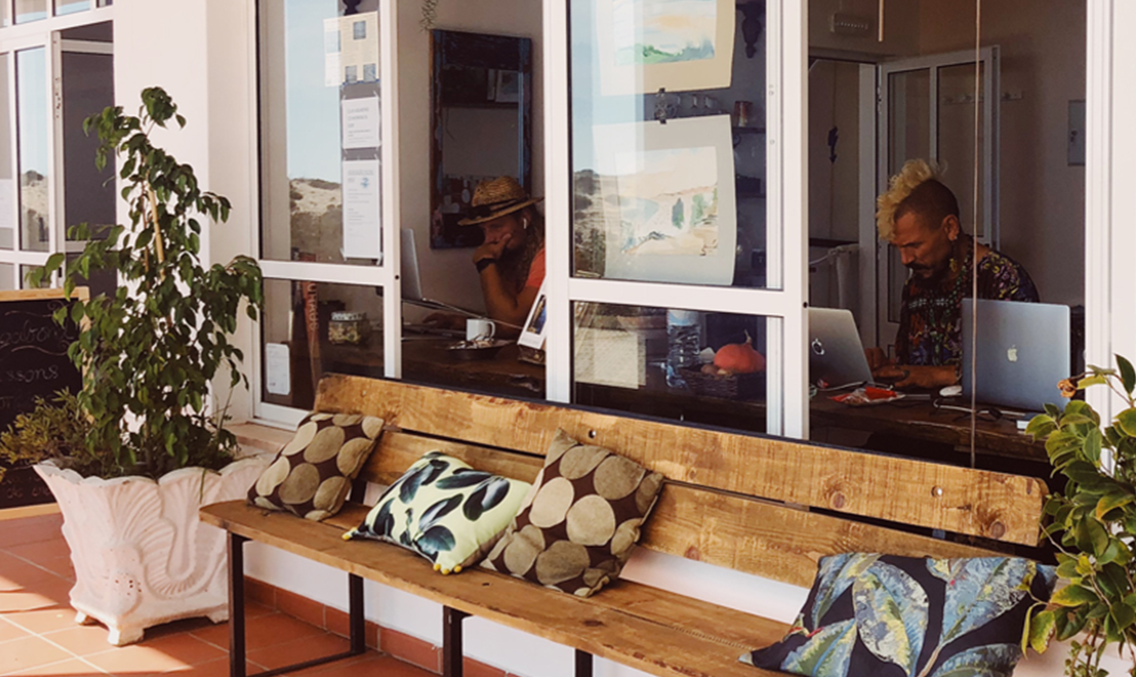 View from outside the Club Vagabond shop showing two coworkers sitting inside, focused on their laptops, working behind open windows.