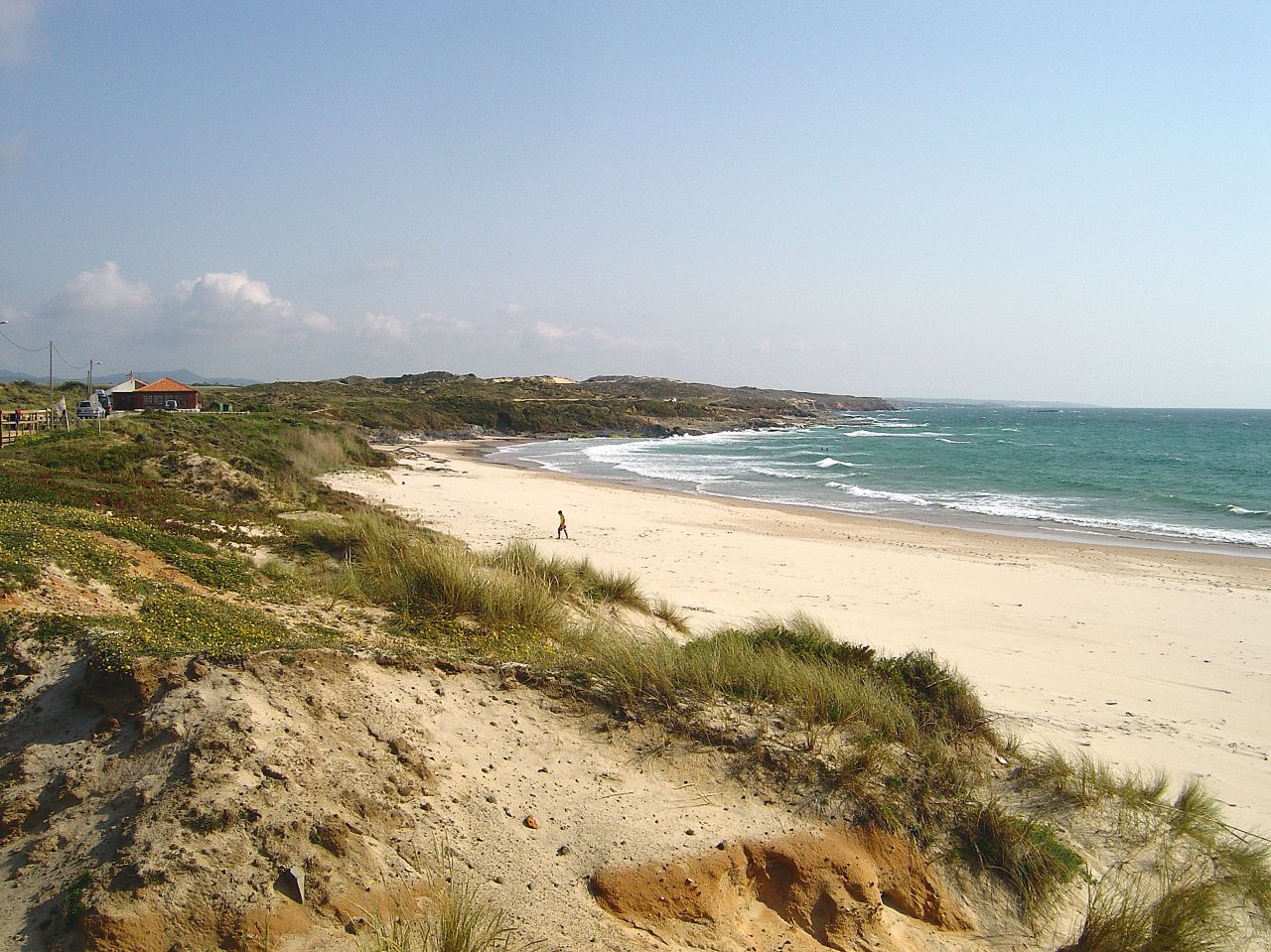 View of São Torpes Beach with turquoise water, gentle waves, a sandy shore, dunes with grass, and a clear blue sky