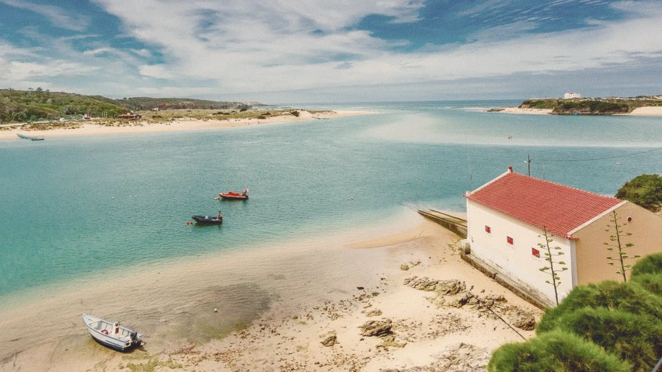 A picturesque view of the Mira River meeting the Atlantic Ocean, with the town of Vila Nova de Milfontes nestled along its banks—both the river and the charming fishing town are visible in the landscape.