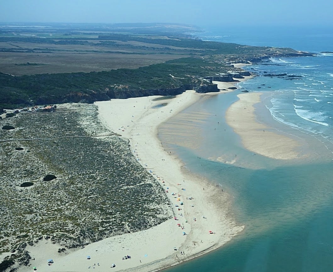 High-angle view of Furnas Beach, featuring a long sandy beach bordered by a rugged coastline with low cliffs covered in trees and bushes